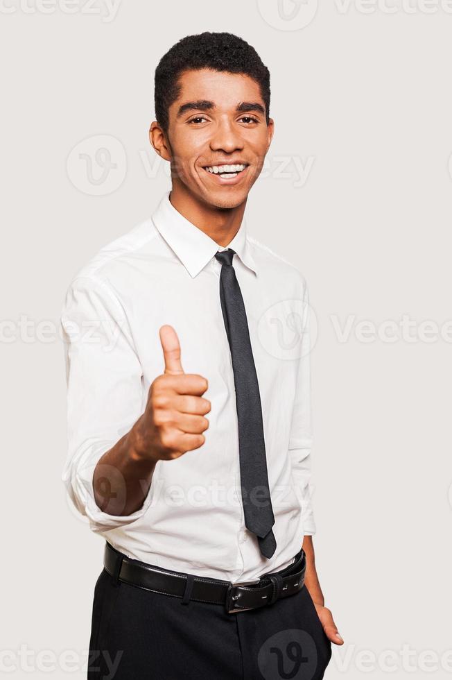 Confident young Afro-American man in formalwear stretching out hand with thumb up while standing against grey background photo