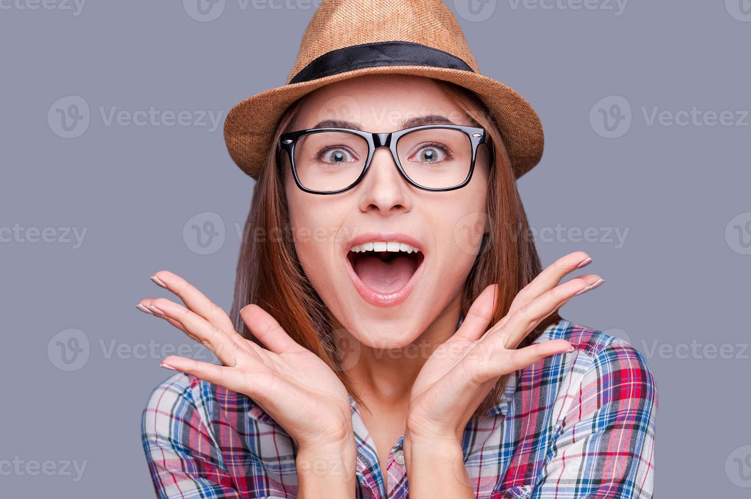 Surprised young woman in glasses and funky hat keeping palms opened looking at camera while standing against grey background photo