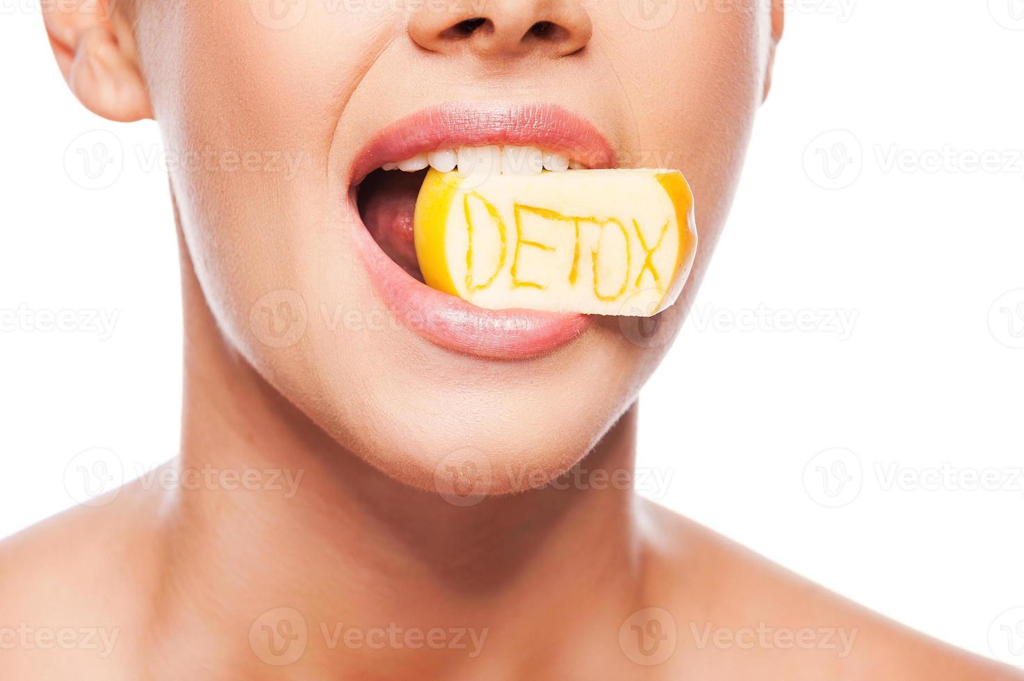Detox diet. Close-up of beautiful young shirtless woman holding piece of apple in her mouth with detox text on it while standing against white background photo