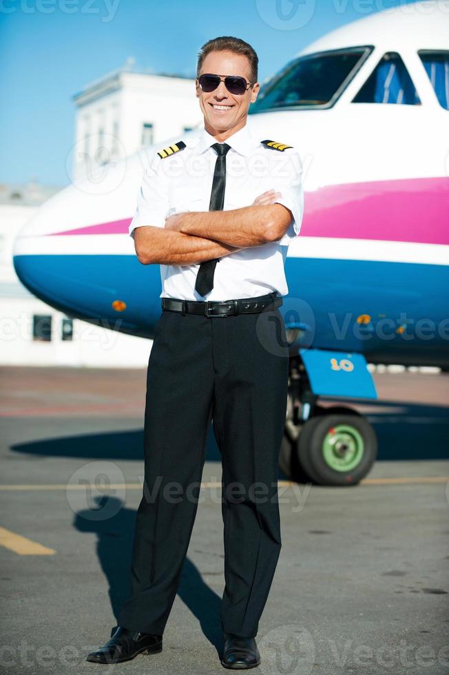 listo para volar. longitud total de piloto masculino confiado en uniforme manteniendo los brazos cruzados y sonriendo con un avión en el fondo foto