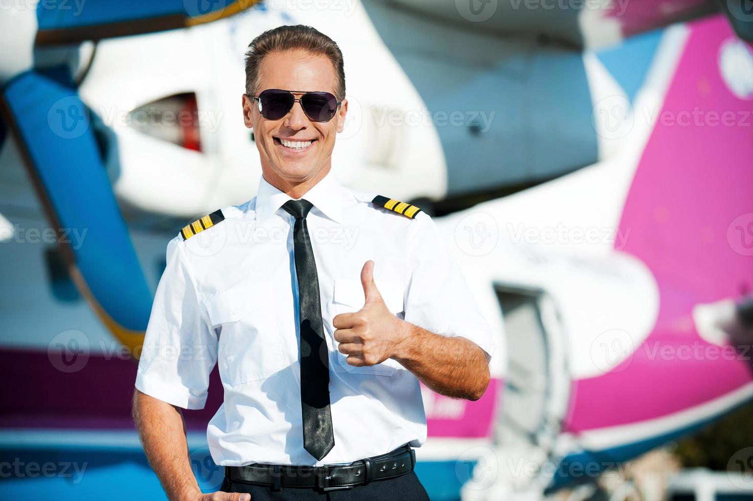 I love my job Confident male pilot in uniform showing his thumb up and smiling while standing in front of the airplane photo
