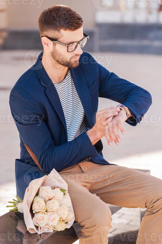 She is late again. Worried young man in smart jacket looking at his watch while sitting on the bench with bouquet of roses laying near him photo