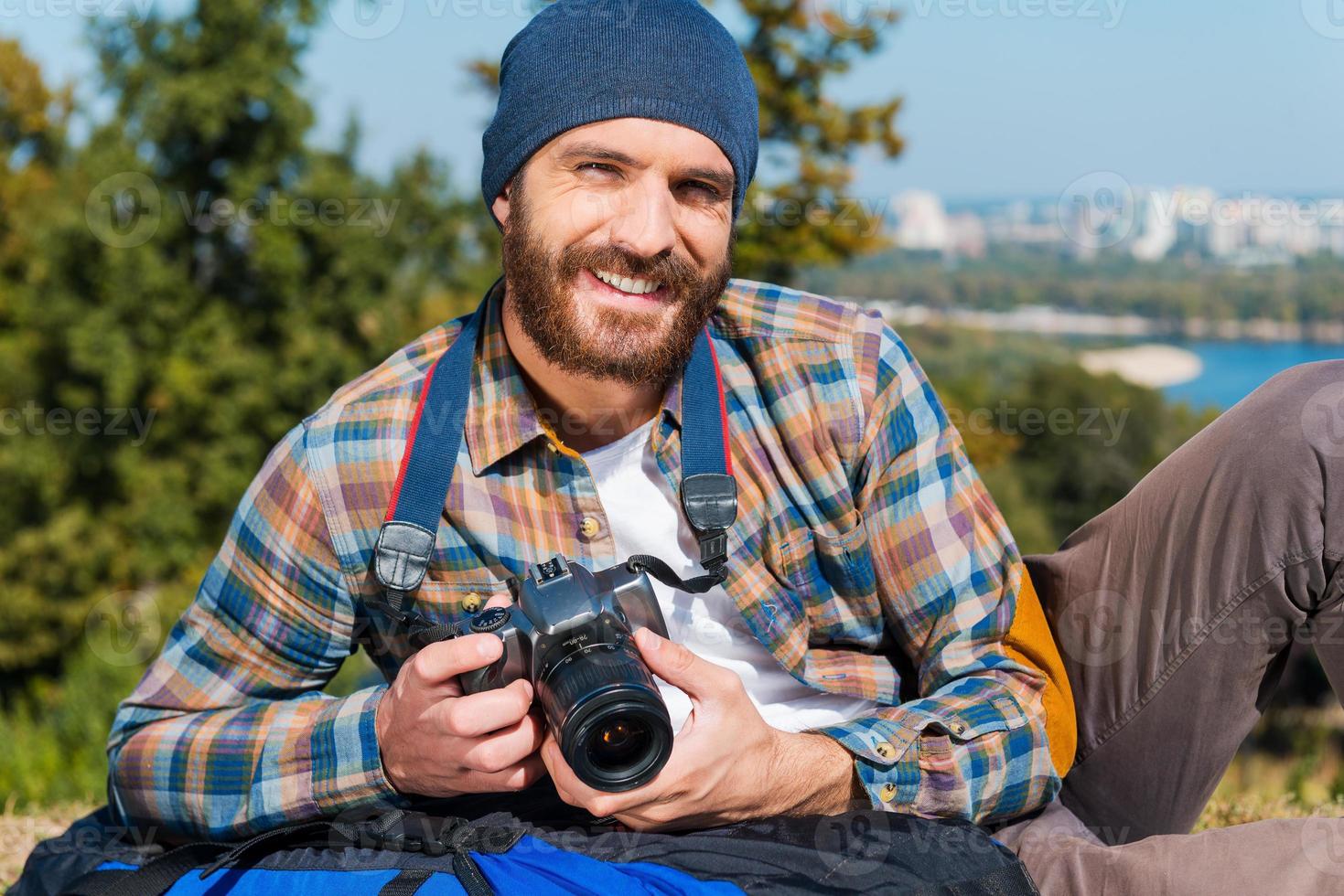 Travelling is impossible without camera.  Handsome young man lying on the backpack and holding camera photo