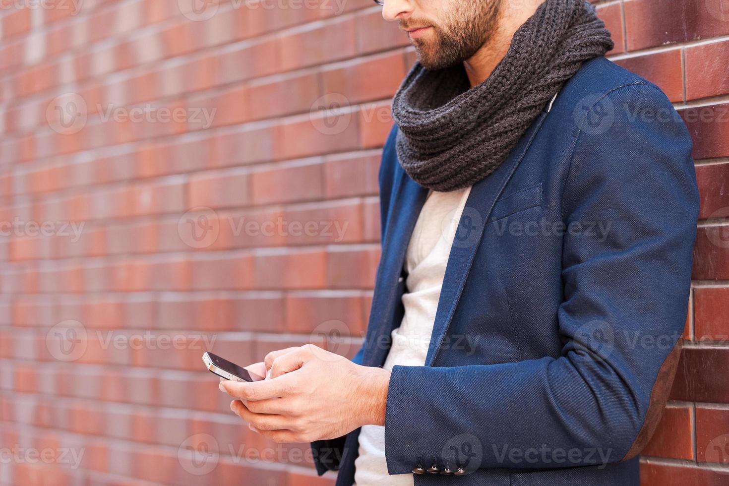 Typing message to friend. Side view of handsome young man in smart casual wear holding mobile phone while leaning at the brick wall photo