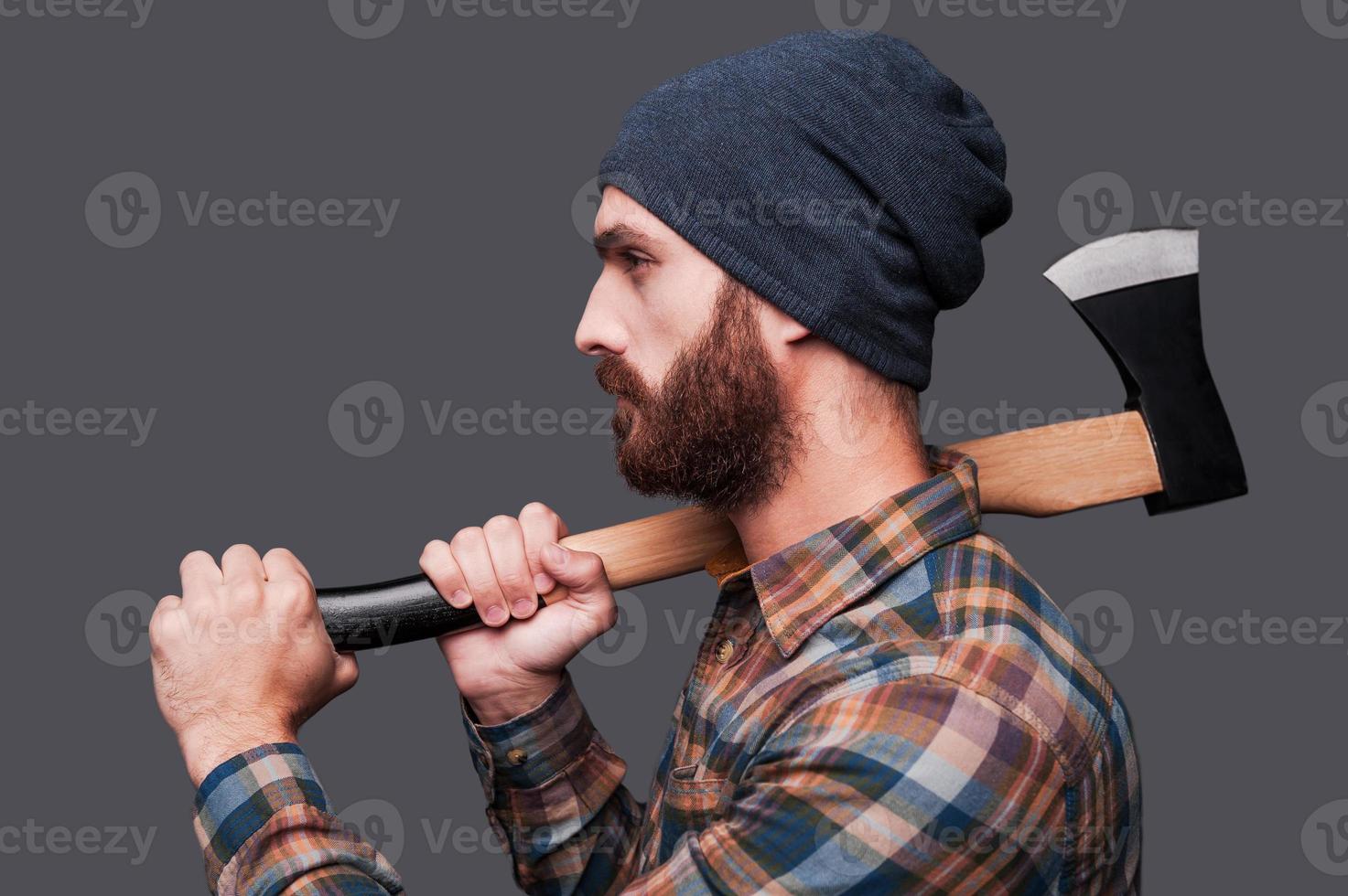 Confident lumberjack. Side view of confident young bearded man holding a big axe and looking at camera while standing against grey background photo