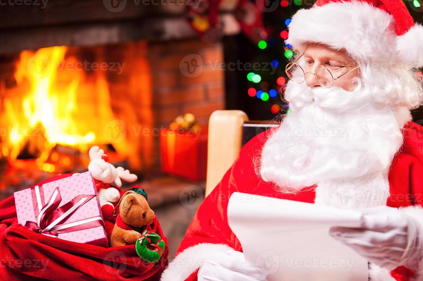 Everyone will get a present. Traditional Santa Claus looking at his sack with presents and holding a paper while sitting at his chair with fireplace and Christmas Tree in the background photo