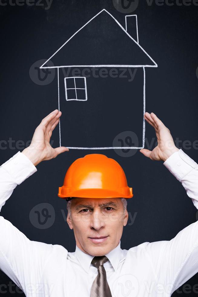 Your house is in the right hands. Confident senior man in formalwear and hardhat holding a chalk drawn house in his hands while standing against blackboard photo