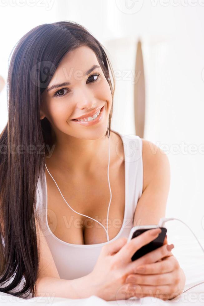 disfrutando de su música favorita. hermosa joven mujer sonriente escuchando el reproductor de mp3 y mirando a la cámara mientras está acostada en la cama foto