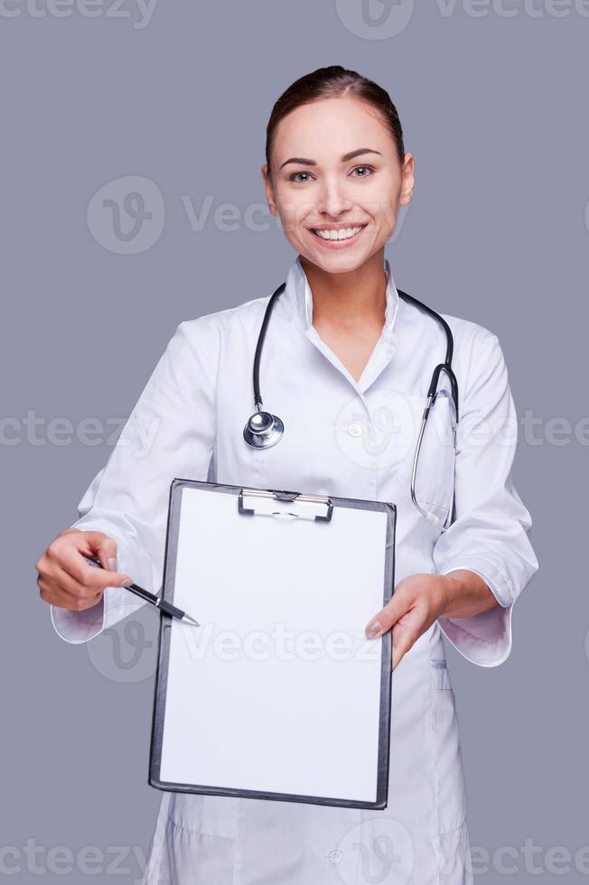 I need your signature here. Confident female doctor in white uniform stretching out clipboard and looking at camera while standing against grey background photo