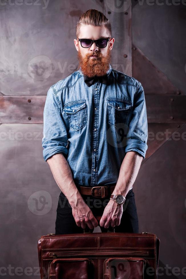 Stylishly confident. Handsome young bearded man in sunglasses looking at camera while holding a suitcase in his hands photo