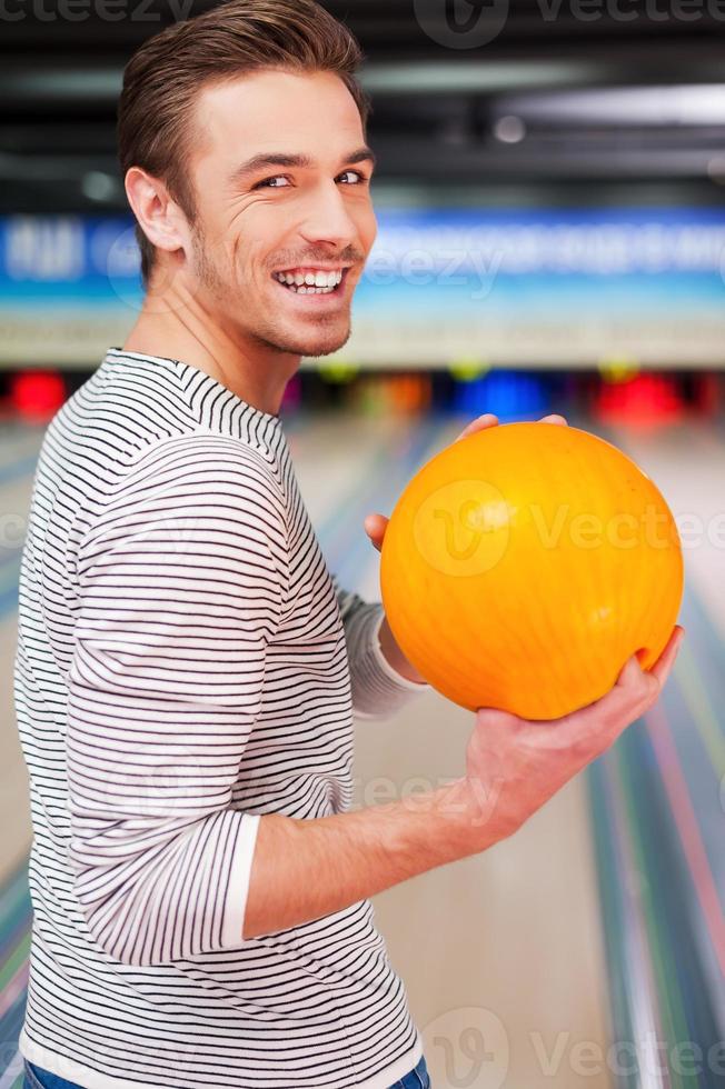 Hoping for a strike. Cheerful young man looking over shoulders and holding a bowling ball while standing against bowling alleys photo