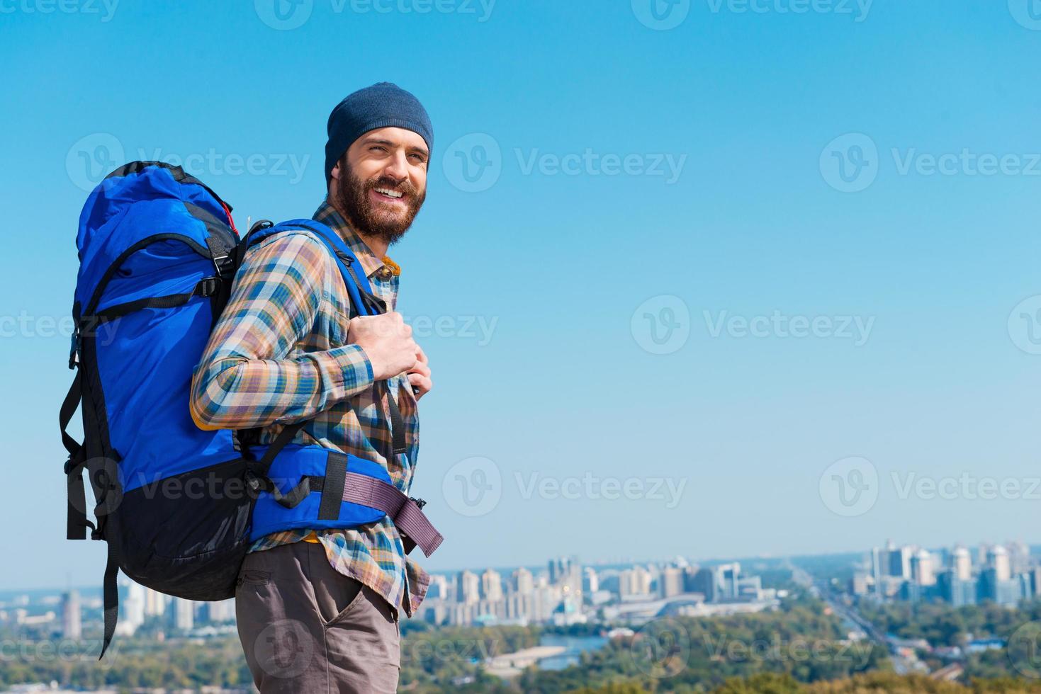 In a search of adventures. Handsome young man carrying backpack and looking at camera through the shoulders a with smile photo