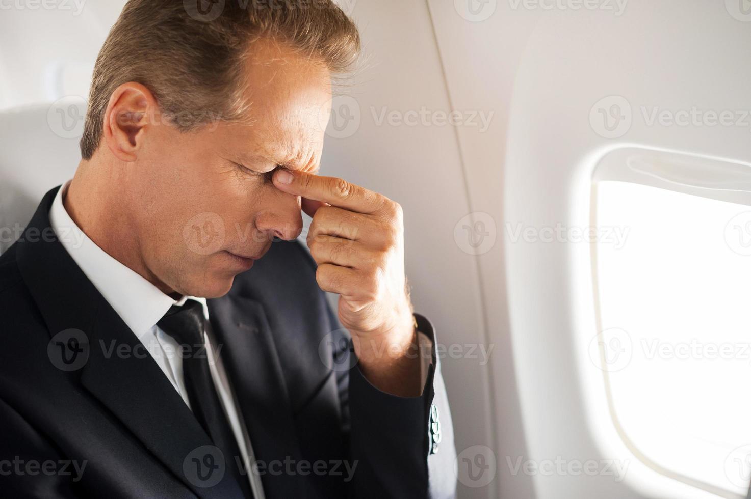 Feeling that awful headache. Frustrated mature businessman touching his face with hand and keeping eyes closed while sitting at his seat in airplane photo