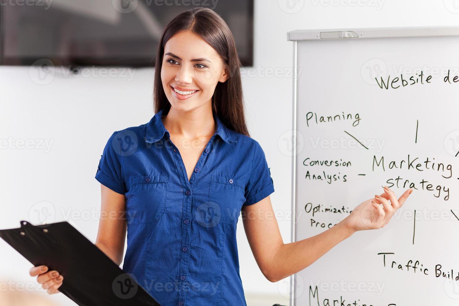 Talking about strategy. Confident young woman in smart casual wear standing near whiteboard and pointing it with smile photo