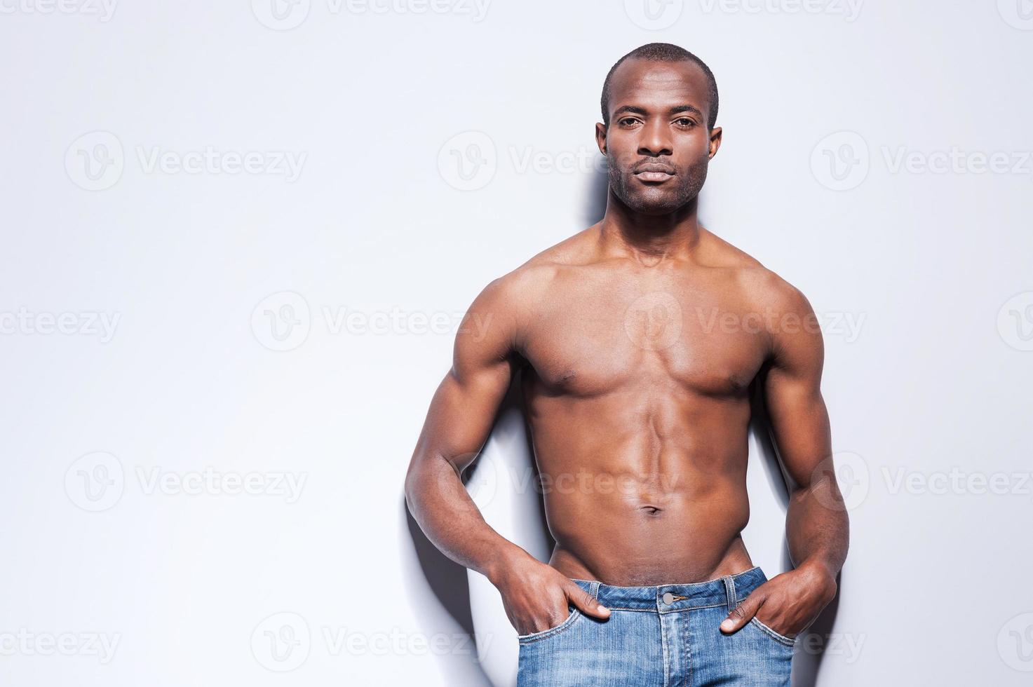 Confident in his perfect body. Handsome young shirtless African man looking at camera and holding hands in pockets while leaning at the grey wall photo