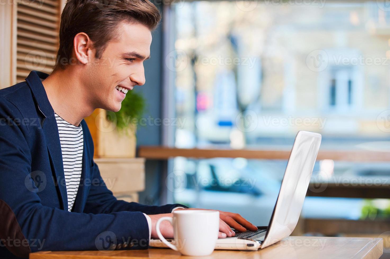 navegando por la red en el café. vista lateral de un joven apuesto que trabaja en una laptop y sonríe mientras disfruta de un café en la cafetería foto