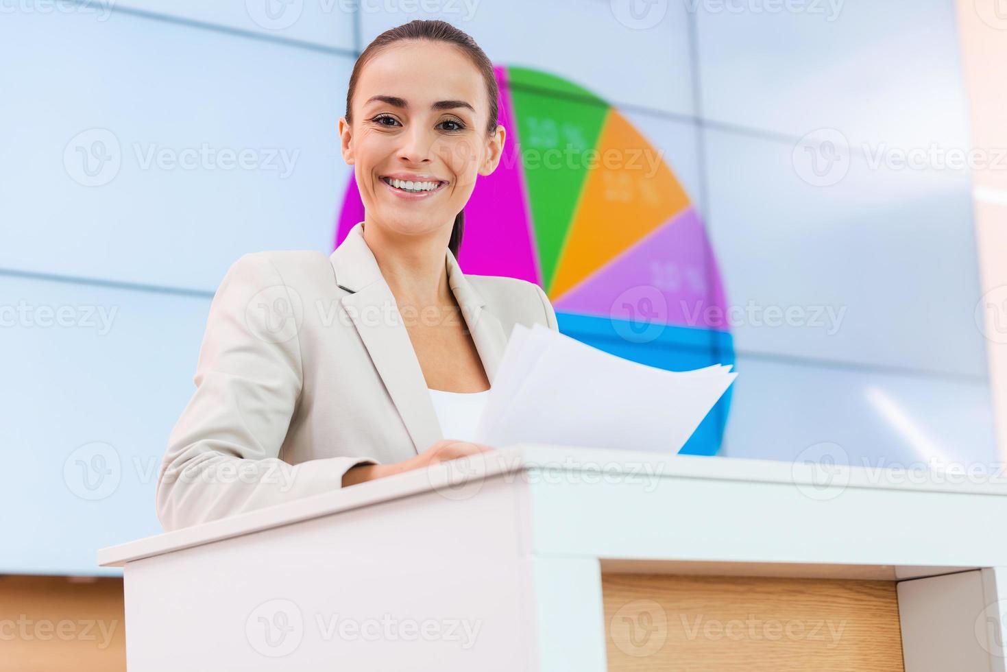 Feeling satisfied with her speech. Low angle view of confident young woman in formalwear standing at the tribune and smiling while making a presentation with projection screen in the background photo