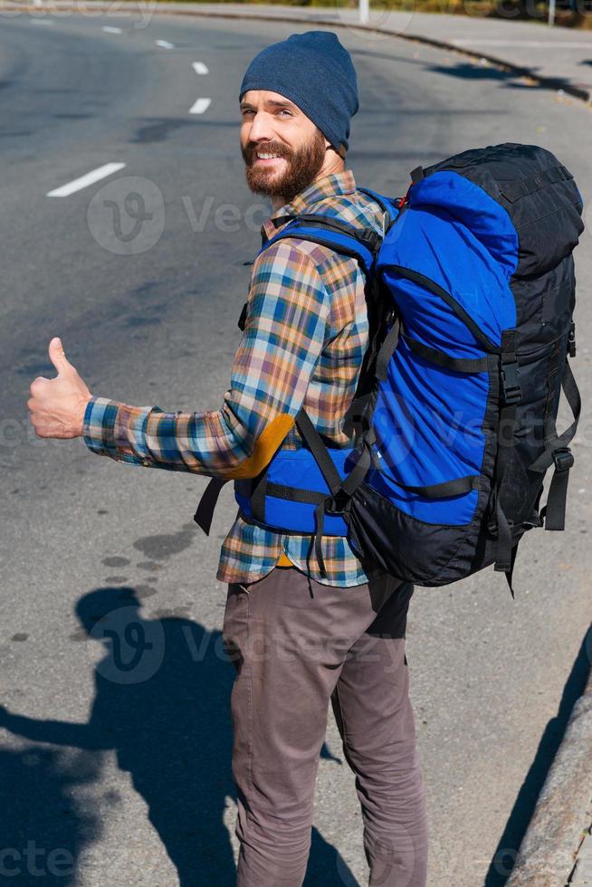 Continuing  the travelling. Handsome young man carrying backpack stretching out hand and looking at camera through the shoulders with thumb up while standing on the road photo