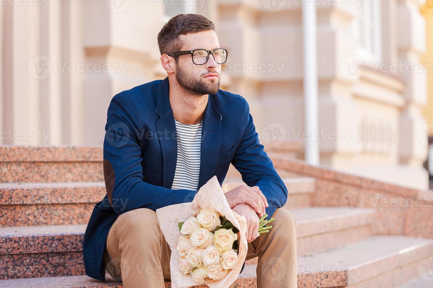 Esperándola. un joven apuesto con una chaqueta inteligente sosteniendo un ramo de flores y mirando hacia otro lado mientras se sienta en la escalera cerca de la casa foto
