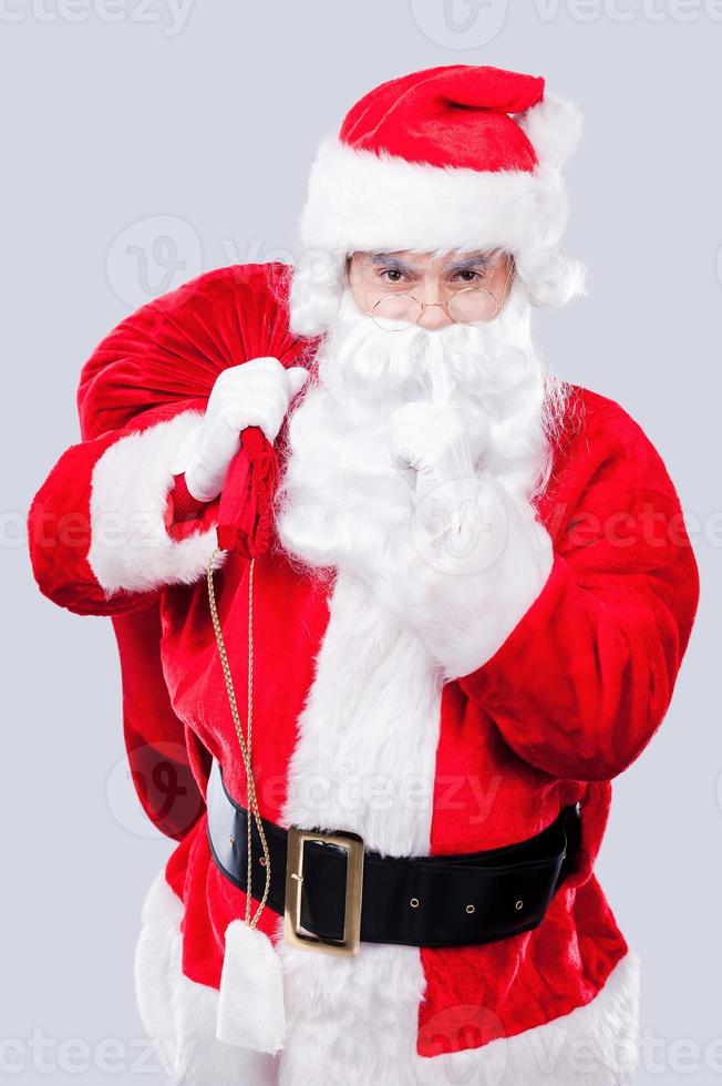 Keep my secret Portrait of traditional Santa Claus holding his finger on lips while carrying sack with presents and standing against grey background photo