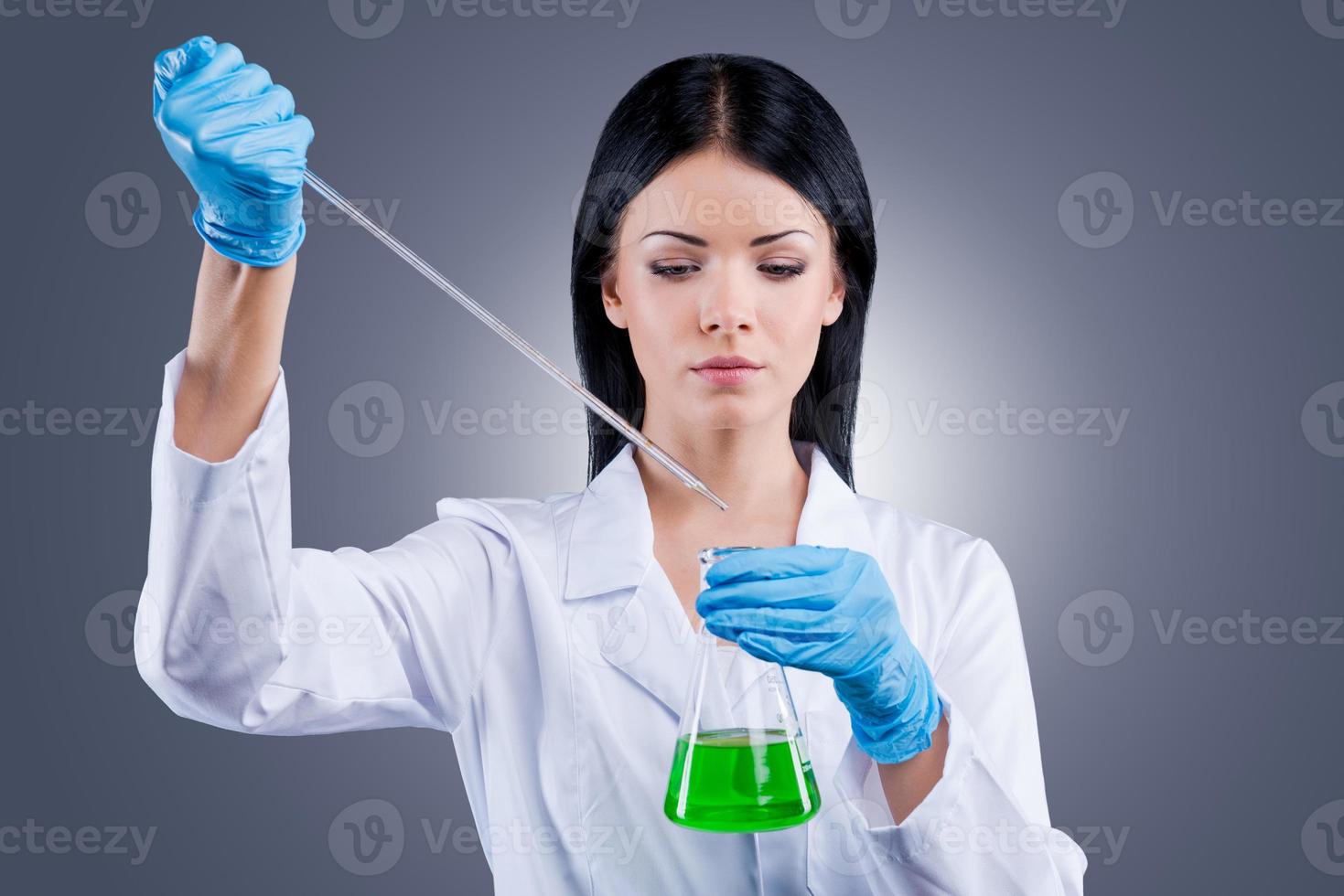 Trying to heal the world. Beautiful female doctor in white uniform holding flasks while standing against grey background photo