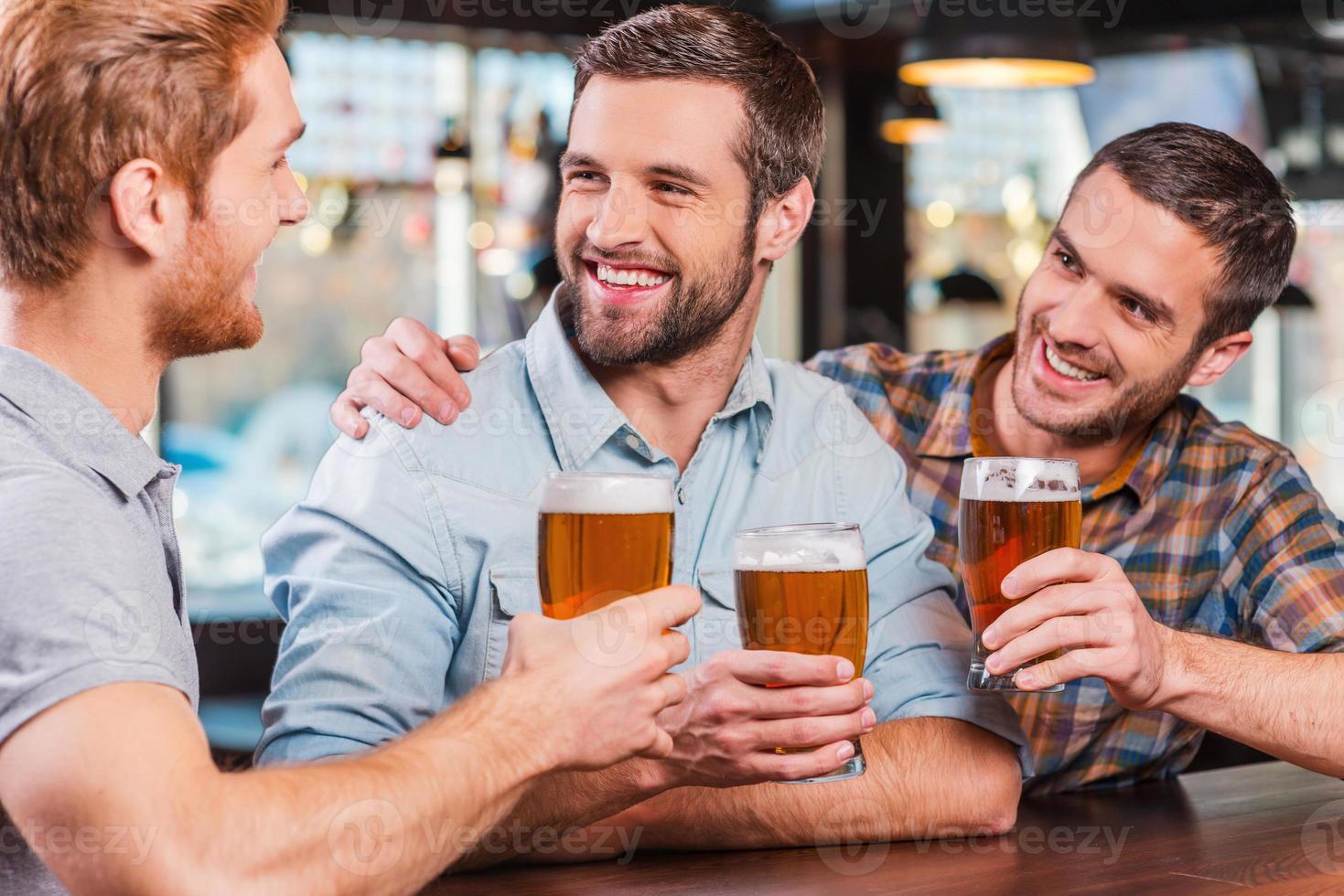 Friends in bar. Three happy young men in casual wear talking and drinking beer while sitting at the bar counter together photo