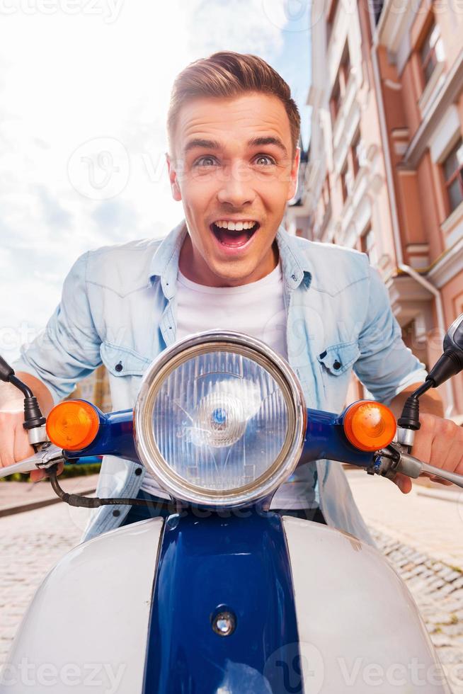 Speed test. Low angle view of happy young man riding scooter along the street and keeping mouth open photo