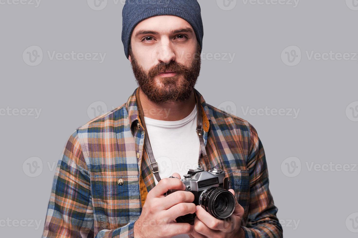 fotógrafo confiado. apuesto joven barbudo sosteniendo una cámara anticuada y mirando la cámara mientras se enfrenta a un fondo gris foto