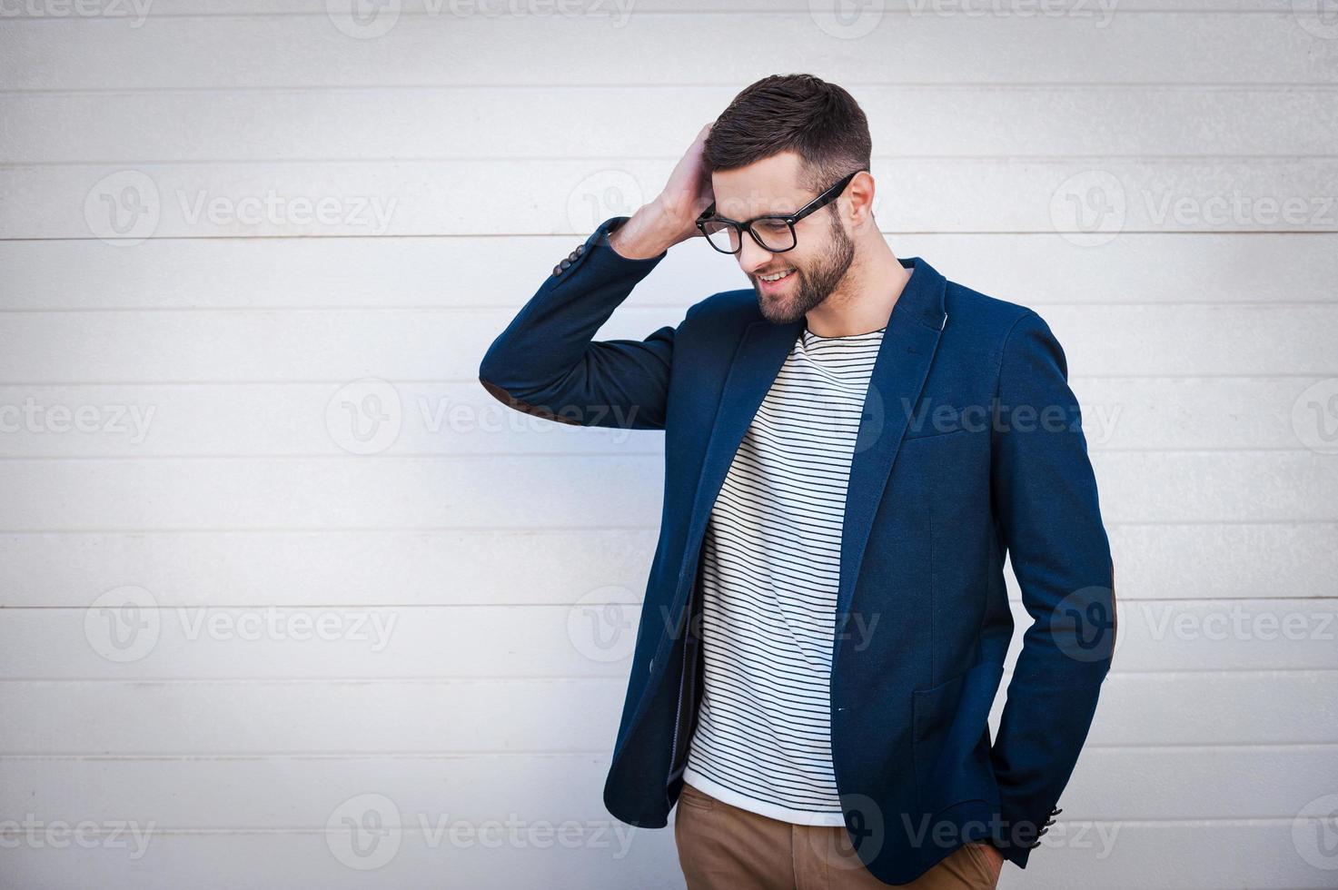 Charming and self-confident. Handsome young man in smart jacket adjusting his hair and smiling while standing in front of the brick wall photo
