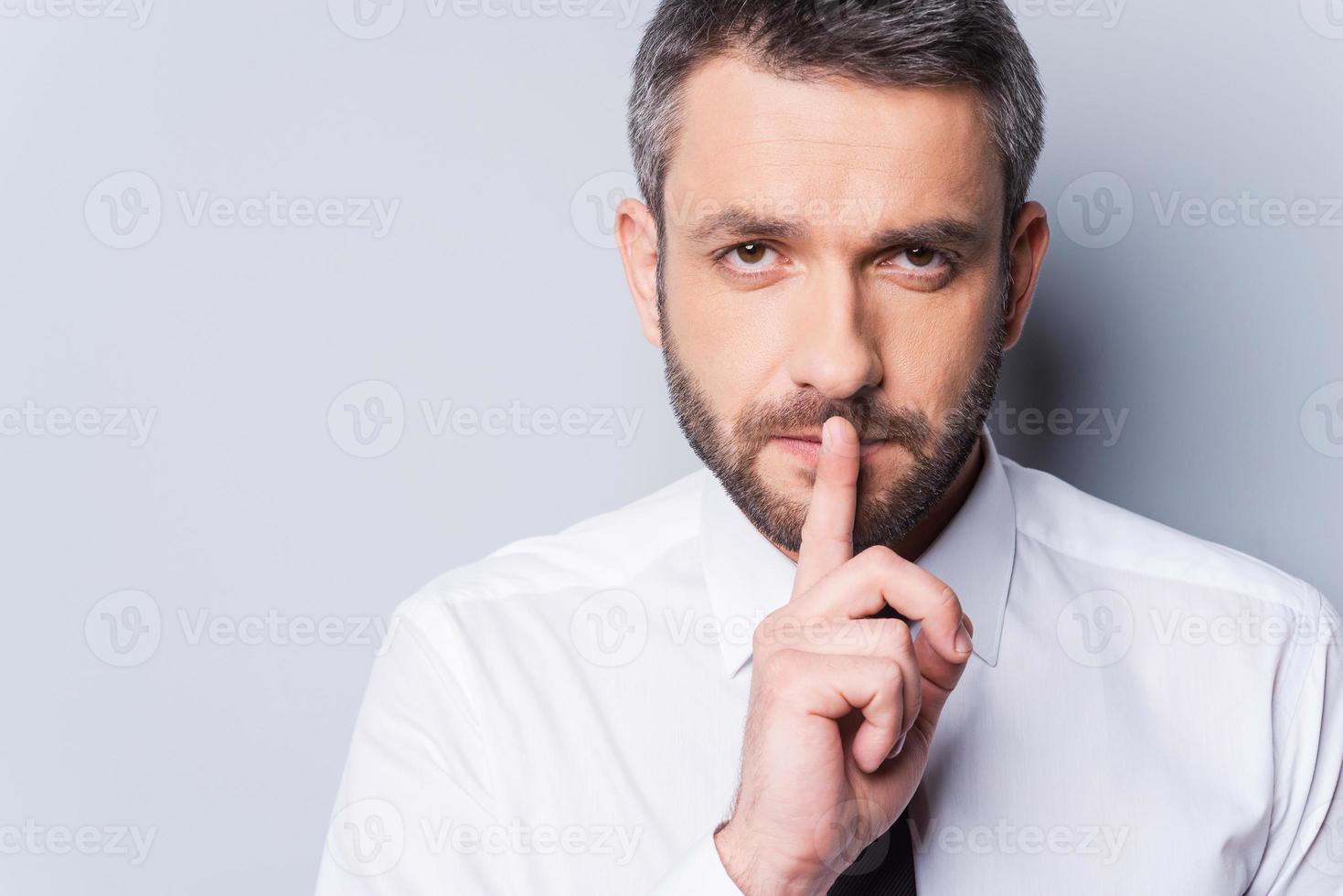 Keep my secret Confident mature man in shirt and tie holding finger on lips and looking at camera while standing against grey background photo