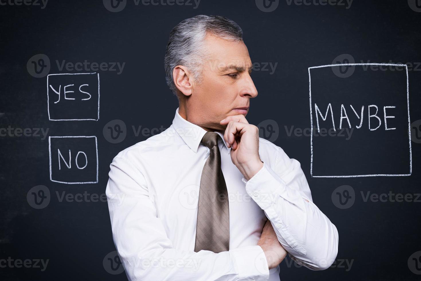 Feeling uncertain. Thoughtful senior man in formalwear holding hand on chin and looking at chalk drawings on blackboard photo