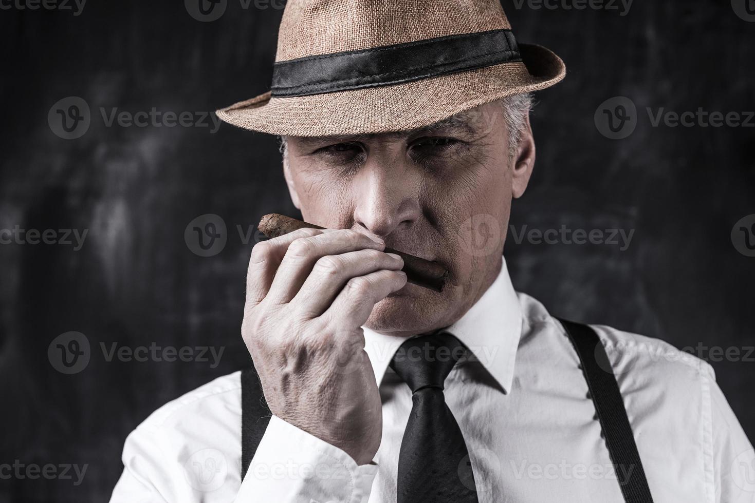 My favorite cigar. Confident senior man in hat and suspenders smelling cigar while standing against dark background photo