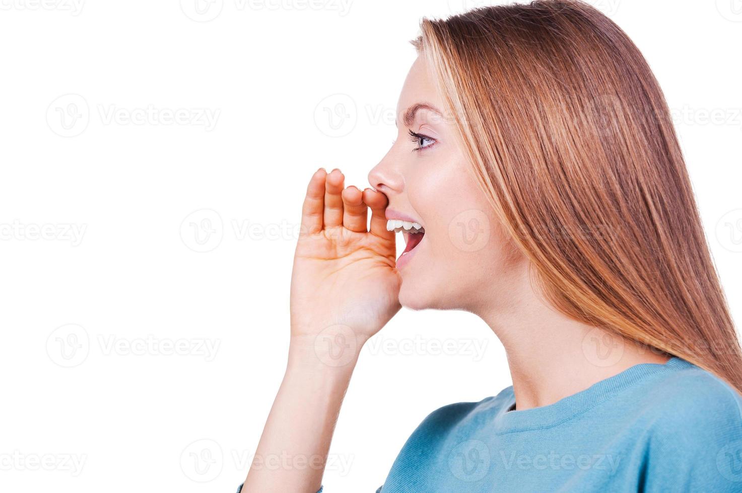 Sharing good news. Side view of beautiful young woman shouting and holding hand near mouth while standing isolated on white photo