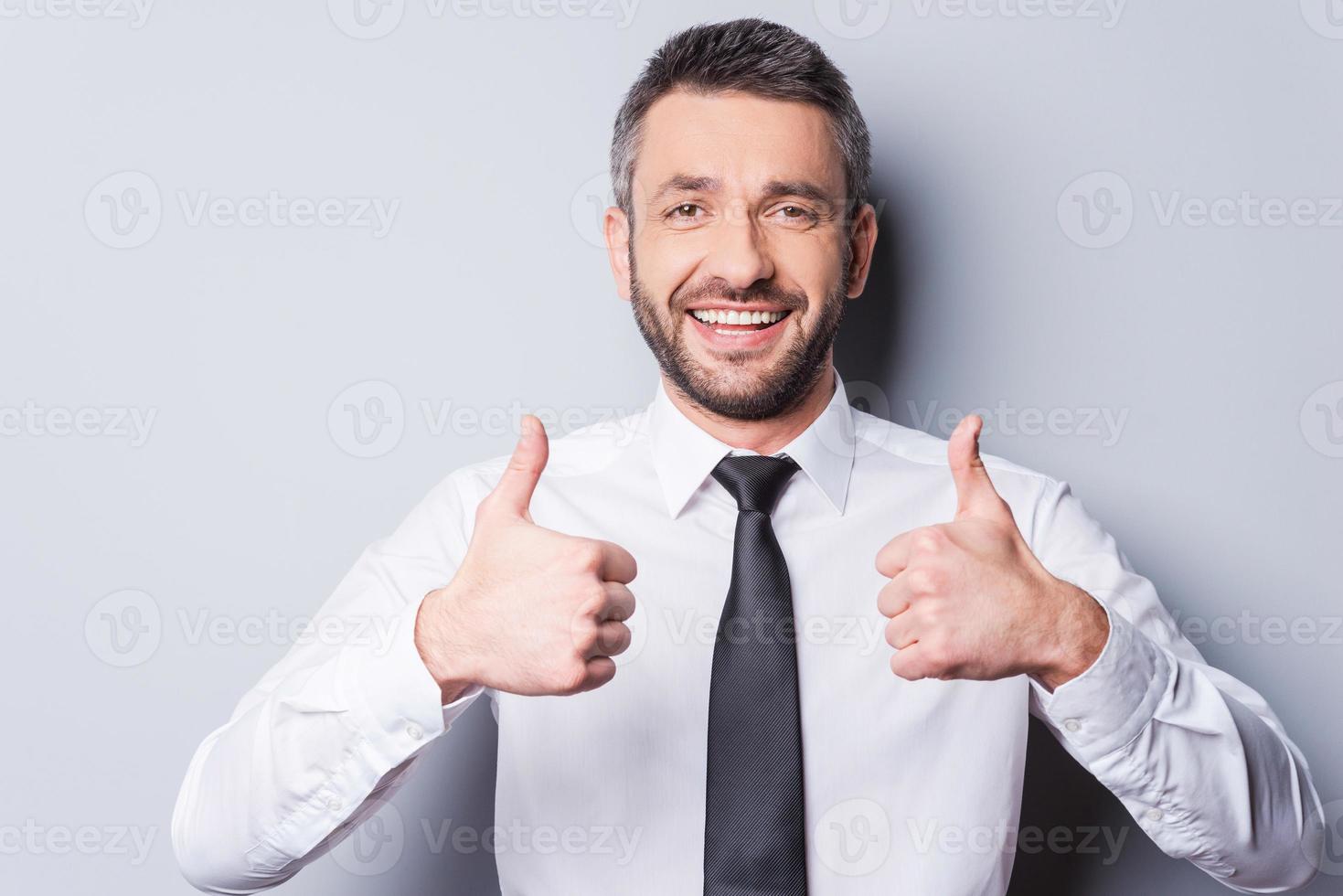 pulgares arriba para el éxito feliz hombre maduro con camisa y corbata mostrando sus pulgares hacia arriba y sonriendo mientras está de pie contra un fondo gris foto