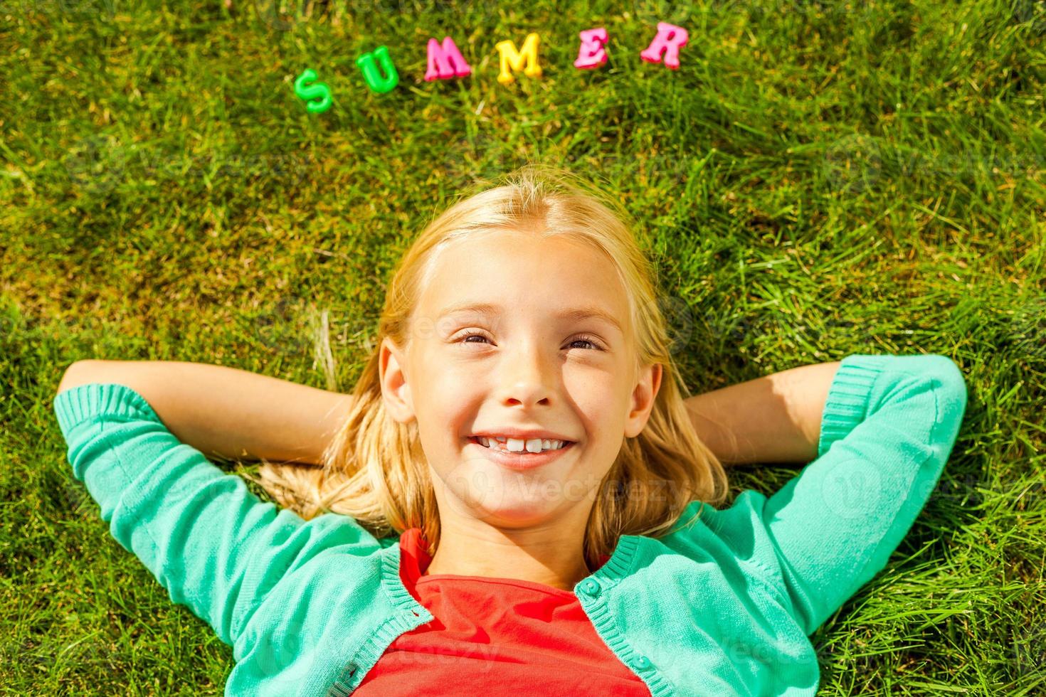 Me encanta la vista superior de verano de una linda niñita cogida de la mano detrás de la cabeza y sonriendo mientras está tumbada en la hierba verde con letras de plástico de colores sobre su cabeza foto