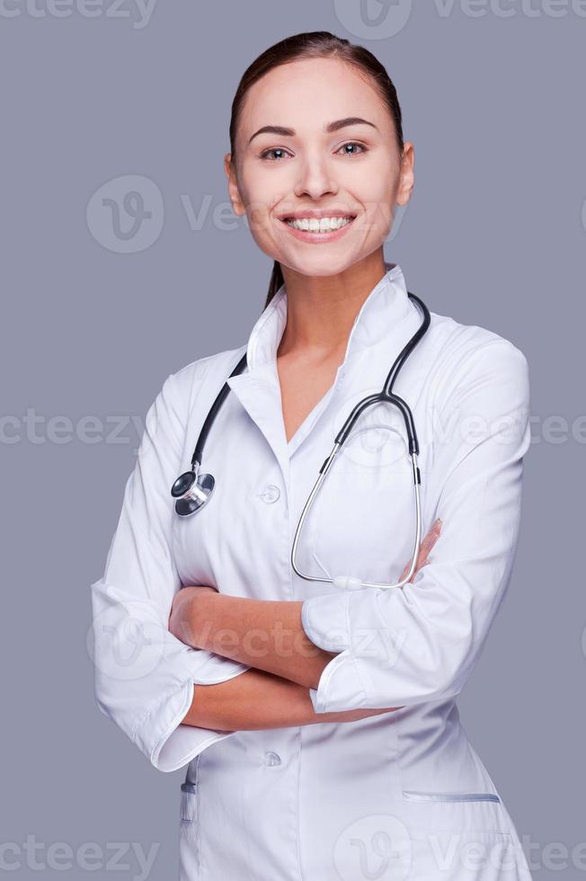 cuidando bien tu salud. doctora segura de sí misma con uniforme blanco mirando a la cámara y sonriendo mientras se enfrenta a un fondo gris foto