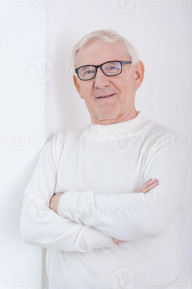 Wise and confidence. Confident senior man keeping arms crossed and smiling at camera while leaning at the wall photo