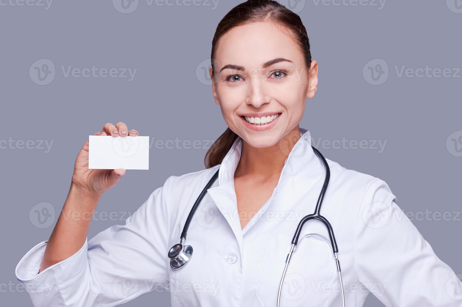 copie el espacio en su tarjeta. doctora confiada en uniforme blanco sosteniendo la tarjeta y mirando a la cámara y sonriendo mientras está de pie contra el fondo gris foto