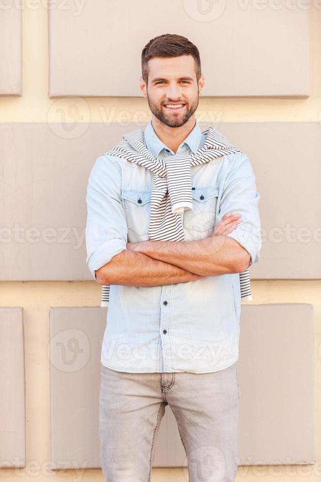 Confident handsome. Handsome young man keeping arms crossed and smiling while standing in front of the textured wall outdoors photo