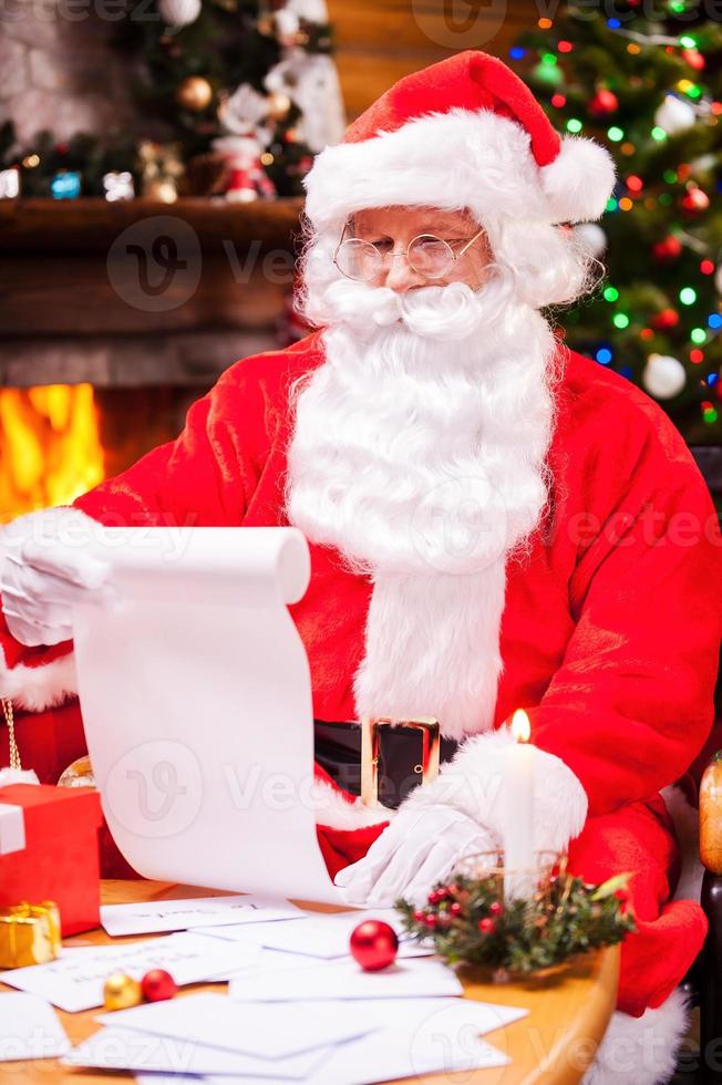 Checking his Christmas list. Concentrated Santa Claus sitting at his chair and reading a letter with envelopes laying on the table photo