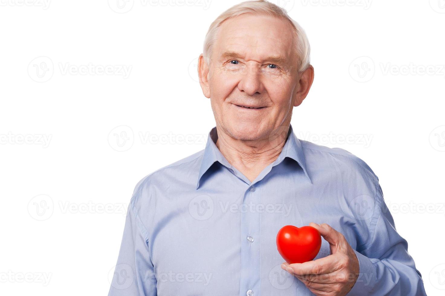 Hope with big heart. Smiling senior man in shirt holding heart prop while standing against white background photo