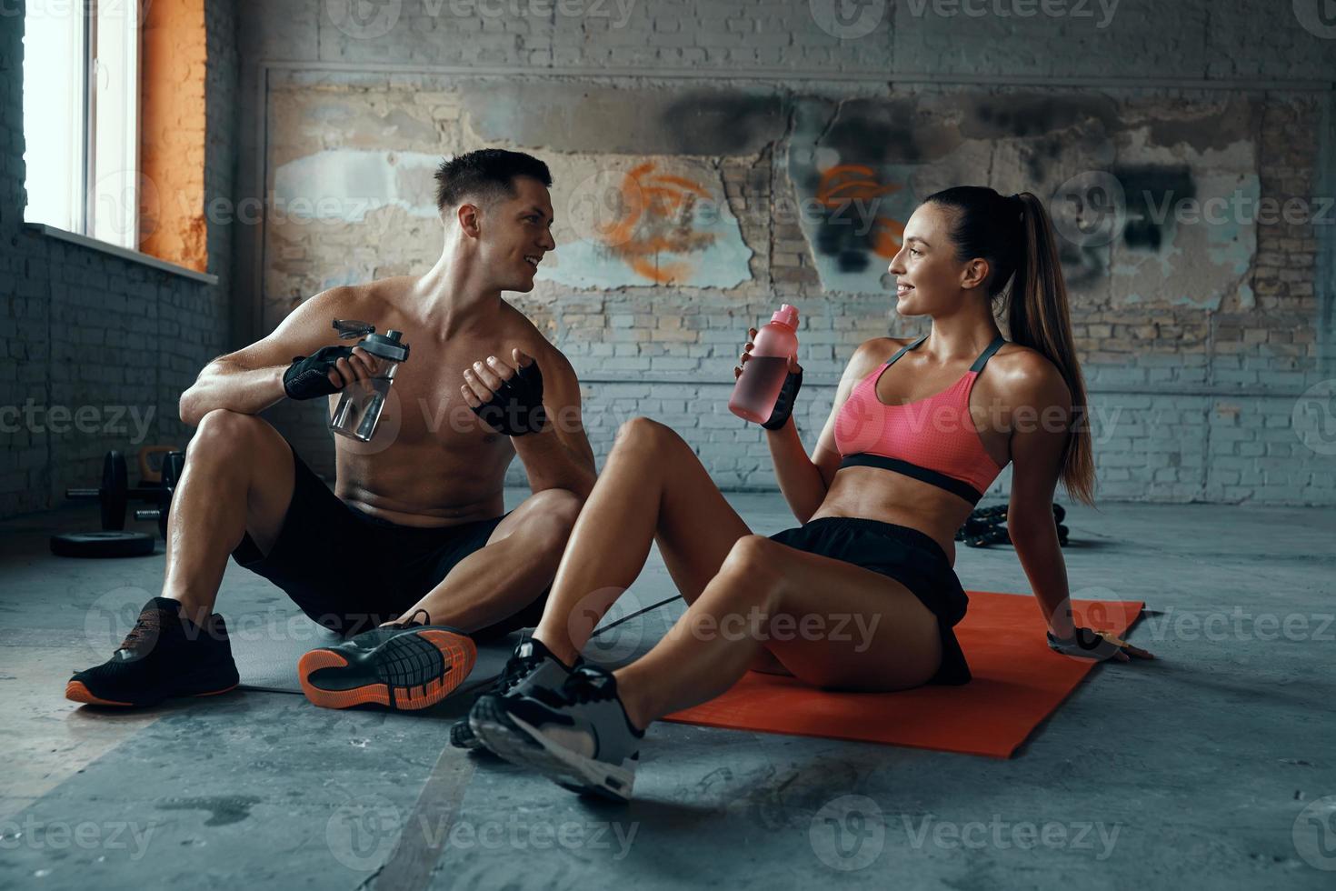 feliz pareja joven bebiendo agua y hablando mientras se sienta en colchonetas de ejercicio en el gimnasio foto