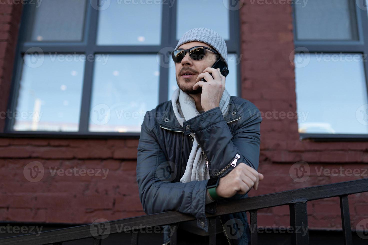 a man in a hat and sunglasses speaks on a mobile phone in front of a brick building with large windows photo