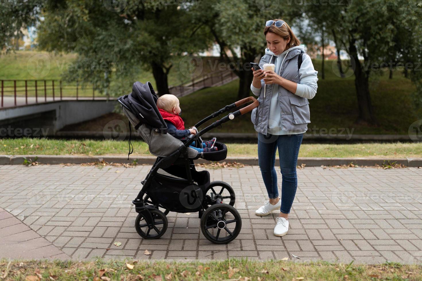 a young mother drinks coffee and watches a mobile phone while walking with a baby in a stroller around the city photo