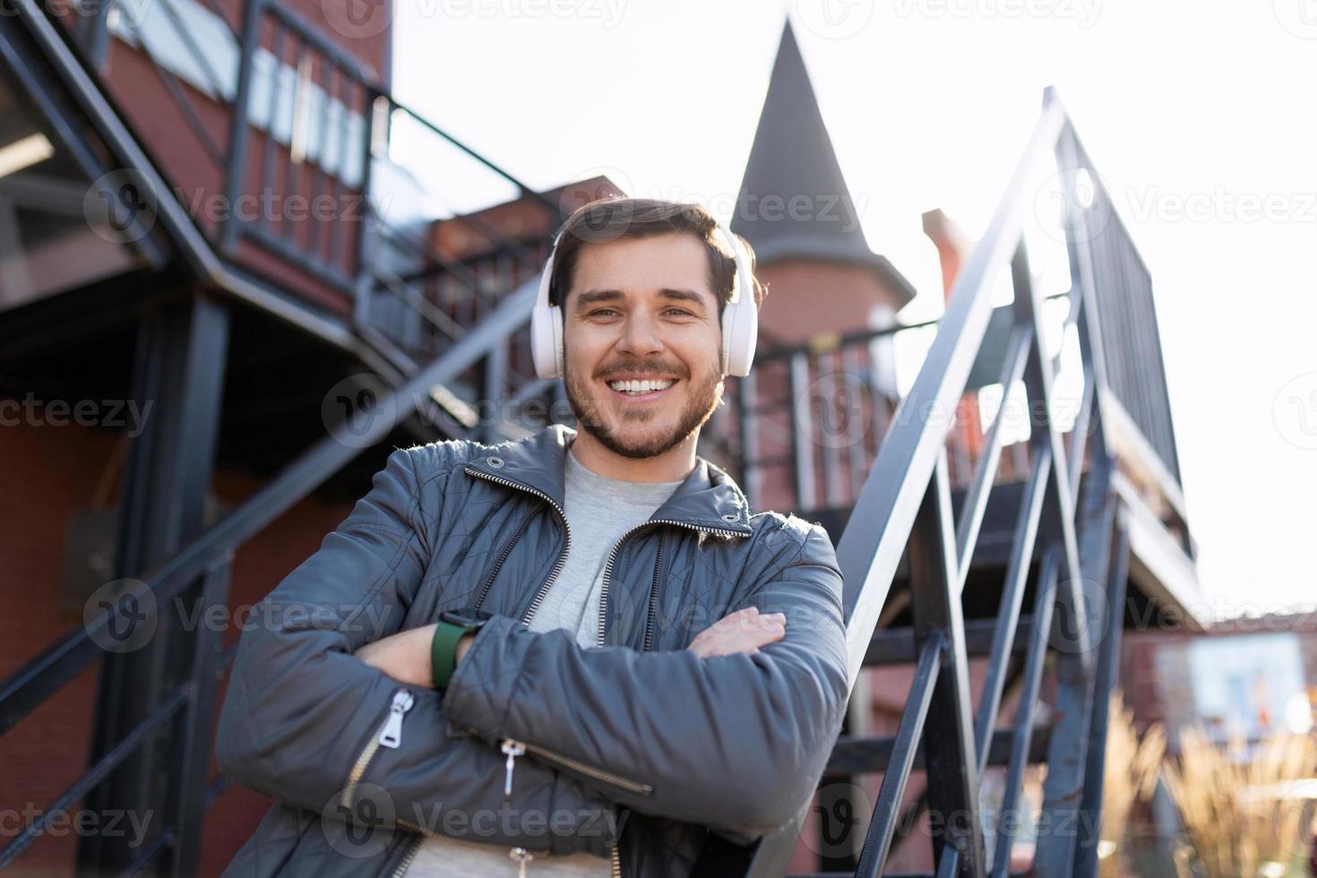 an adult man with a broad smile on his face listens to music in headphones with his arms crossed on his chest against the backdrop of stairs outside the office photo