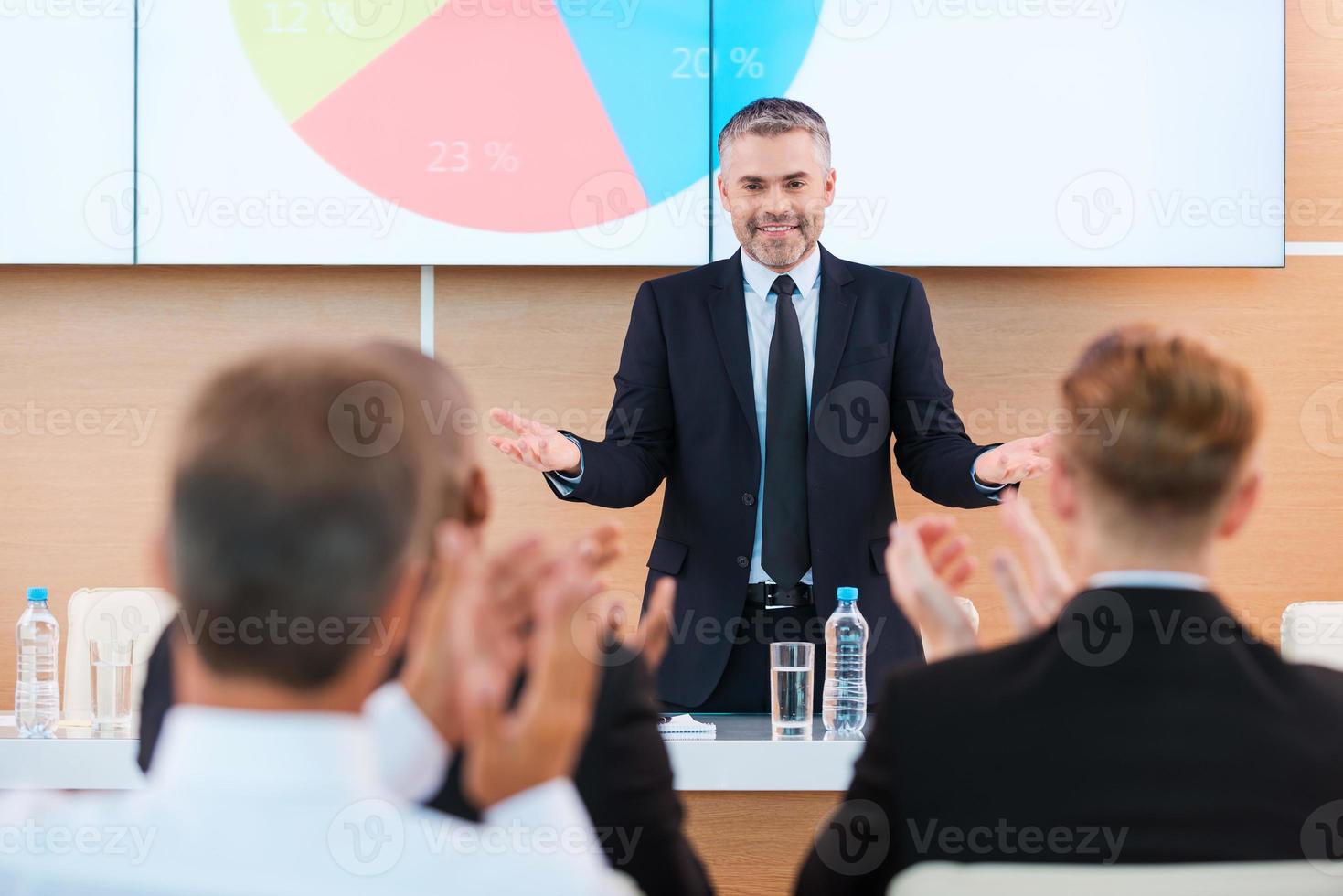 Celebrating success. Confident mature man in formalwear gesturing and smiling while making presentation in conference hall with people applauding on the foreground photo