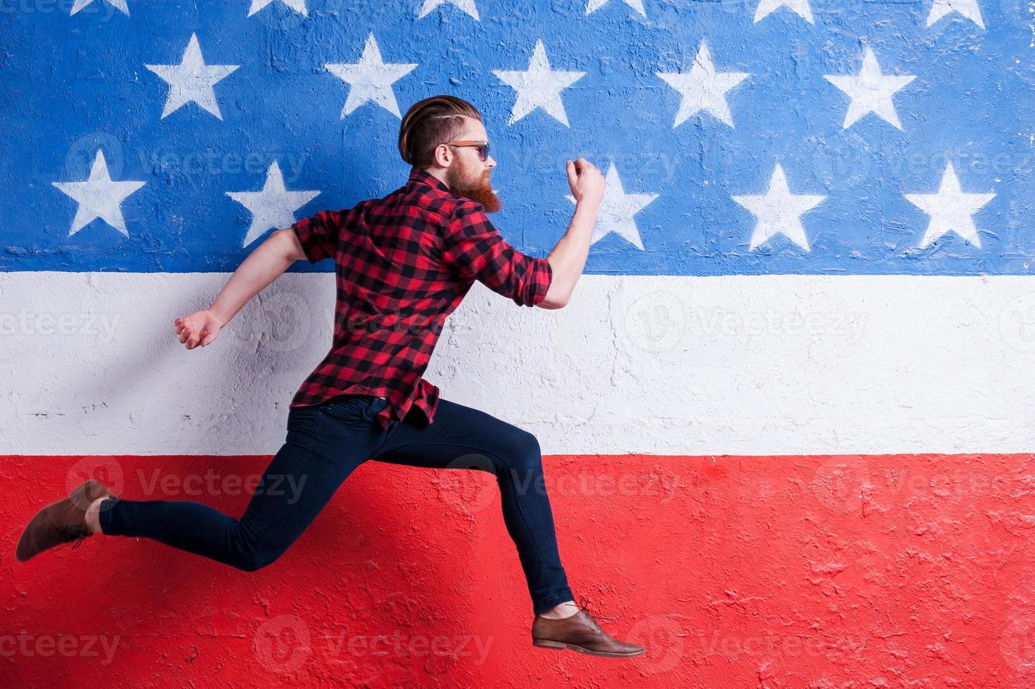 Hurrying to be in trend. Handsome young bearded man wearing sunglasses and running along American flag photo