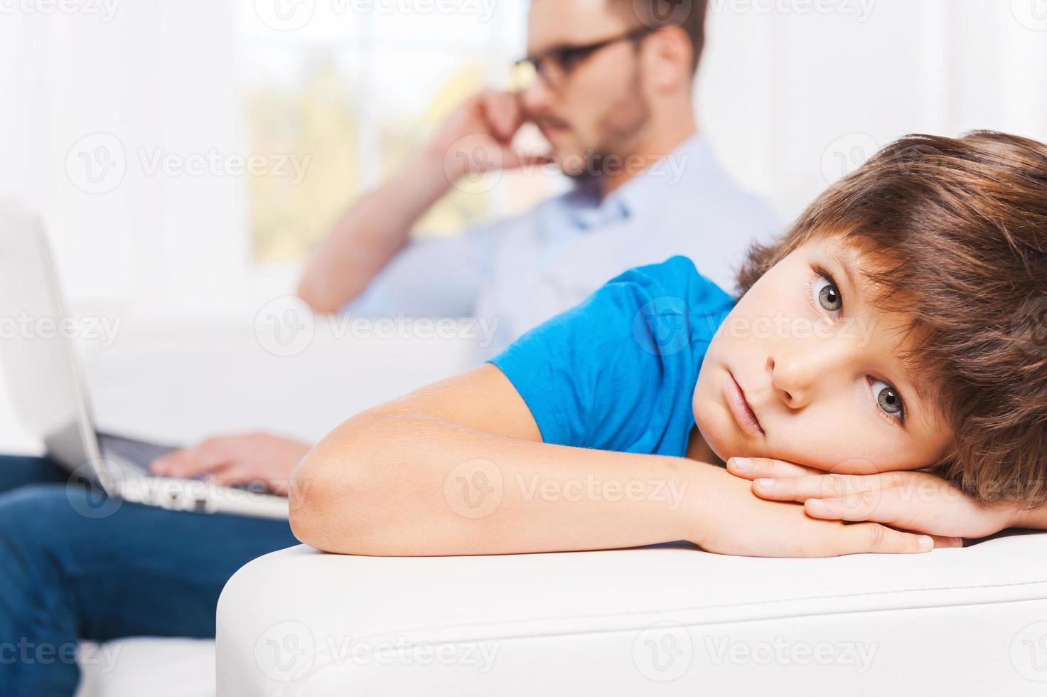 Why he always ignoring me Busy young man working on laptop and talking on the mobile phone with his son sitting on foreground and looking upset photo
