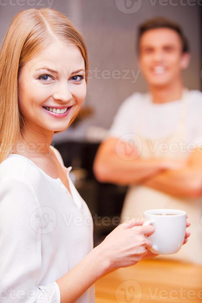 cliente feliz hermosa joven sosteniendo una taza de café y sonriendo mientras está de pie cerca del mostrador del bar y con un alegre barista de pie en el fondo foto