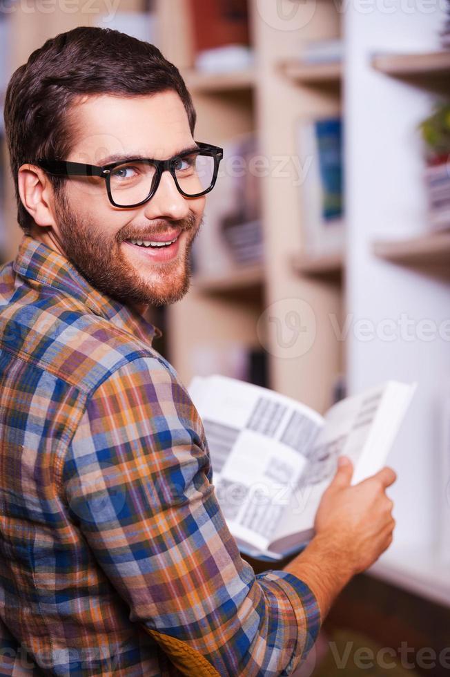 Handsome bookworm. Rear view of cheerful young man holding book and looking over shoulder while standing in front of the bookshelf photo
